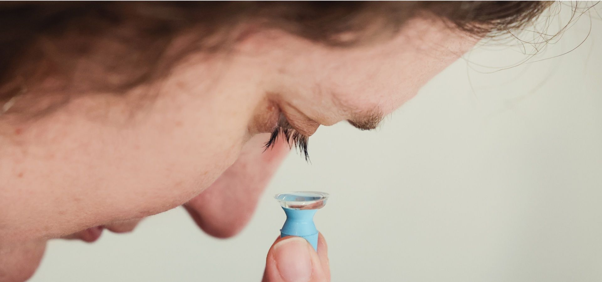Person holding acscleral lens, about to insert it into their eye. Close-up, neutral background.