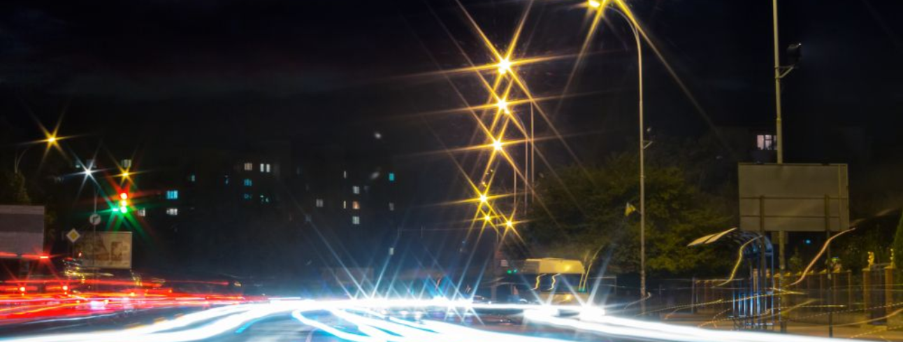 Nighttime road with streaking car lights under a row of bright streetlights against a dark sky.