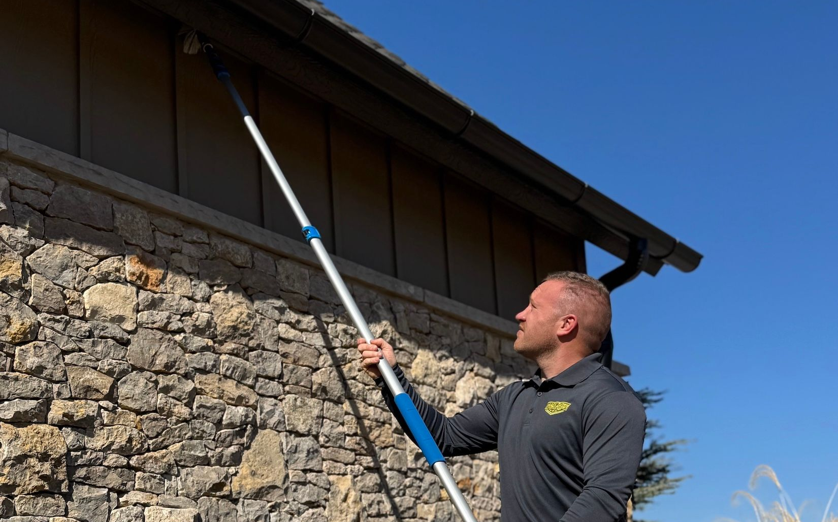 Man cleaning a house exterior with an extendable pole; brown siding, stone wall, clear sky.