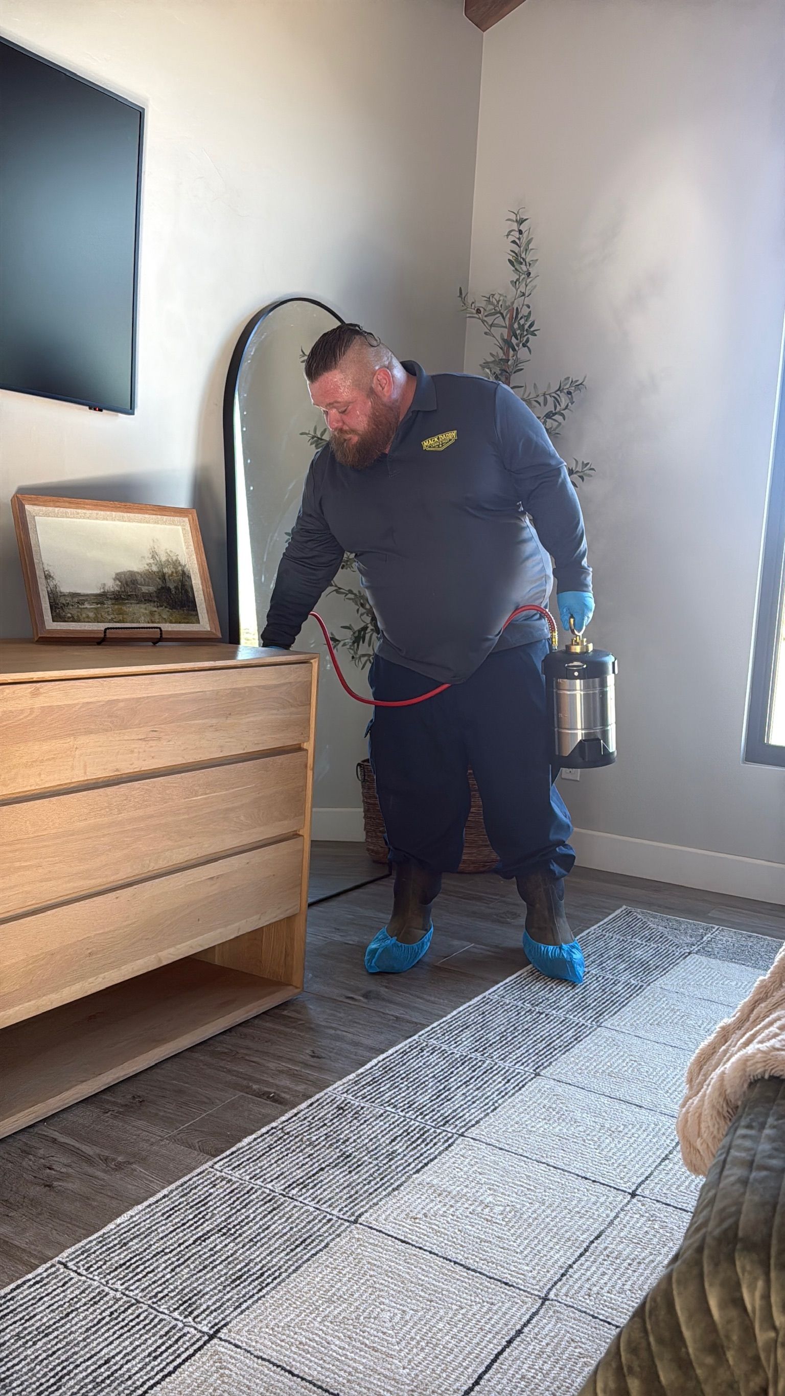 Pest control worker spraying a room. He wears protective gear, and sprays around a dresser and rug.