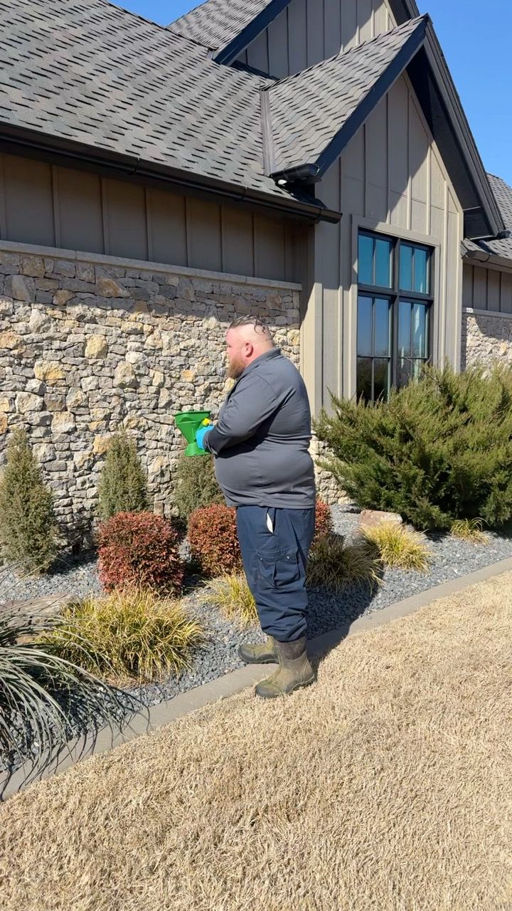 Man sprays greenery near a stone-walled house.