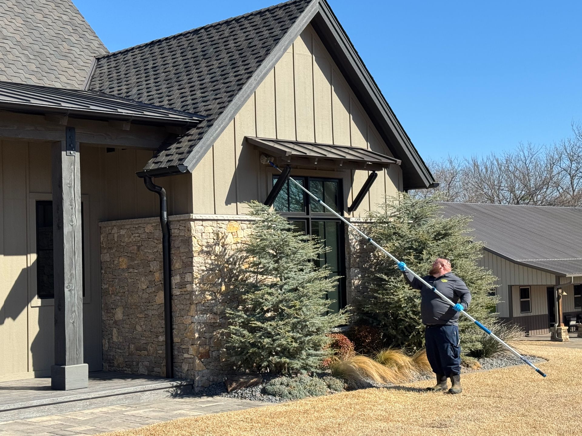 Man using a long tool to clean a window of a house on a sunny day.
