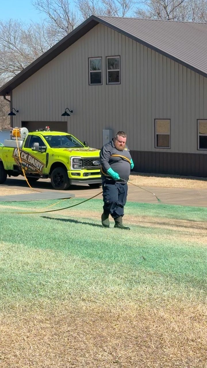 Man spraying lawn with a neon yellow truck in front of a tan building.
