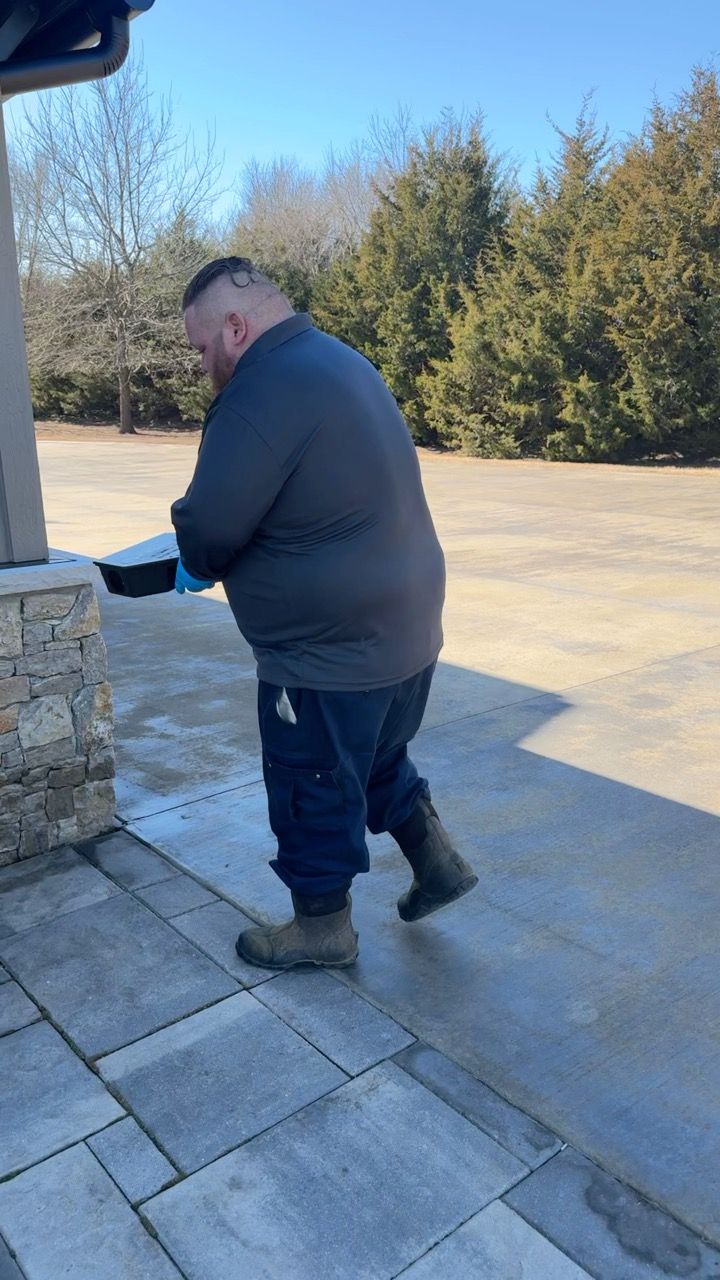 Man walking outside, holding a black box. Wearing blue shirt, pants, and work boots. Sunny day.