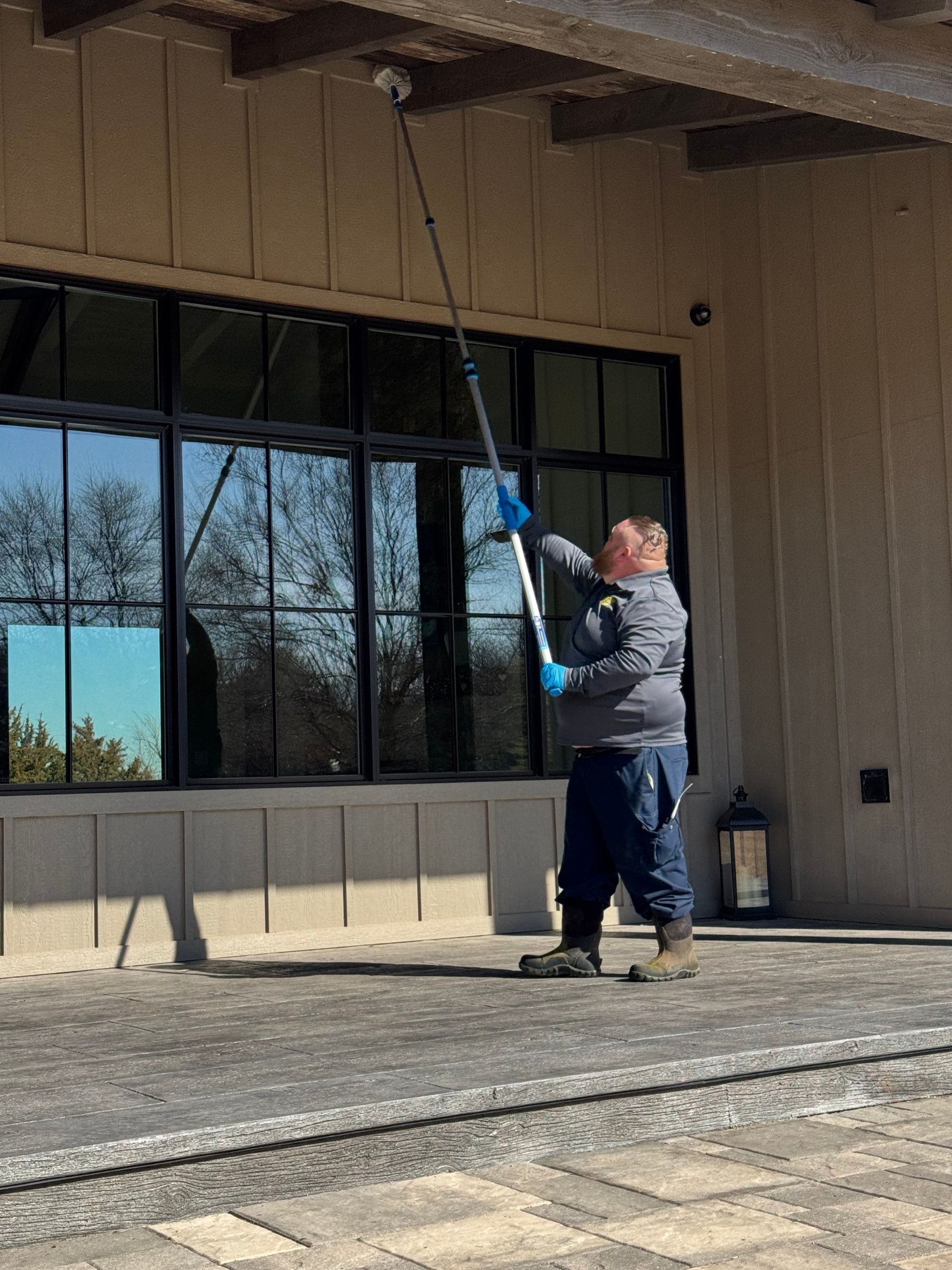 Man using a long-handled tool to clean the underside of a building overhang. Exterior setting with windows.