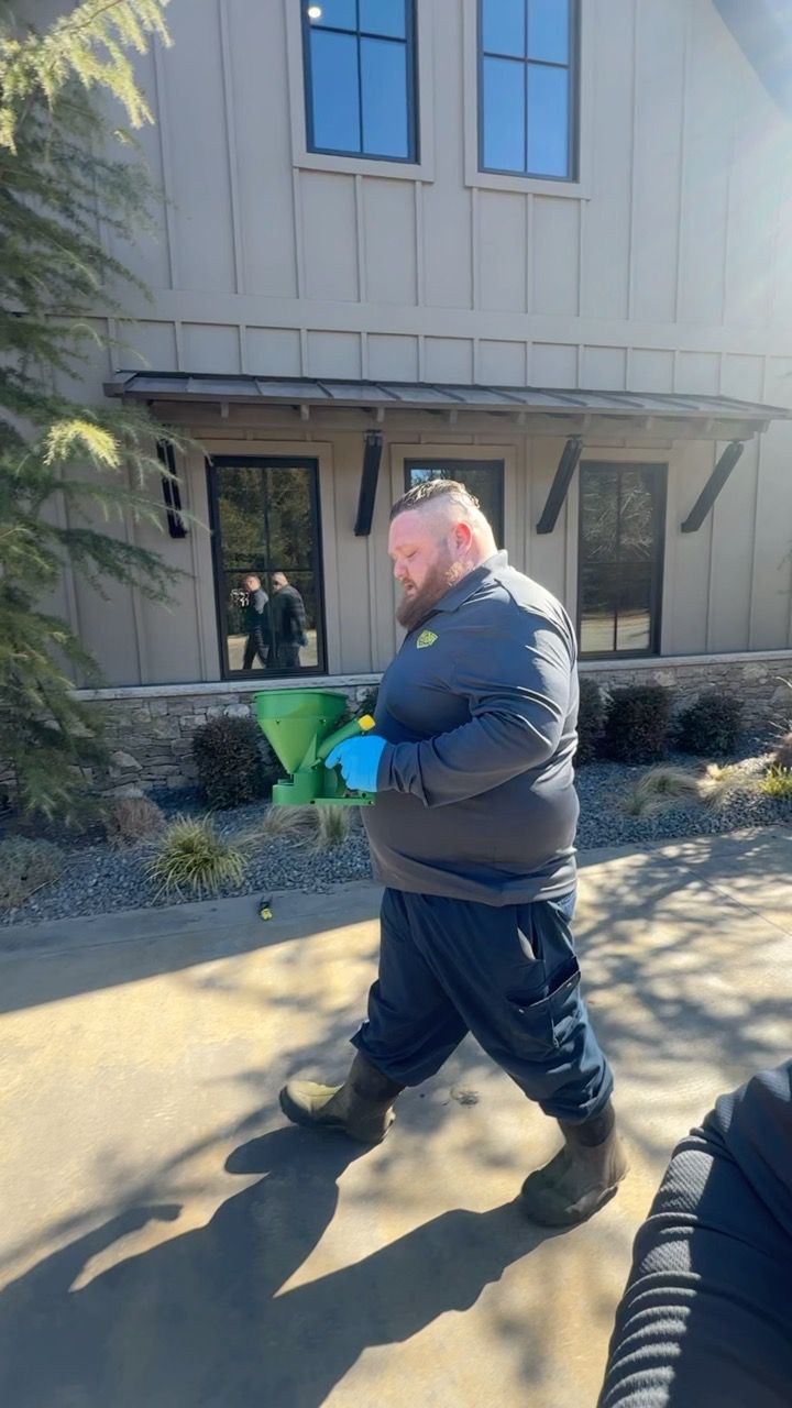 Man in work clothes carrying a green container near a building with windows. Sunny day.