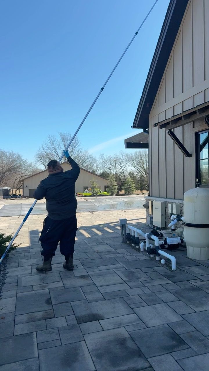 Man uses a long pole to clean a building's exterior near pool equipment on a sunny day.