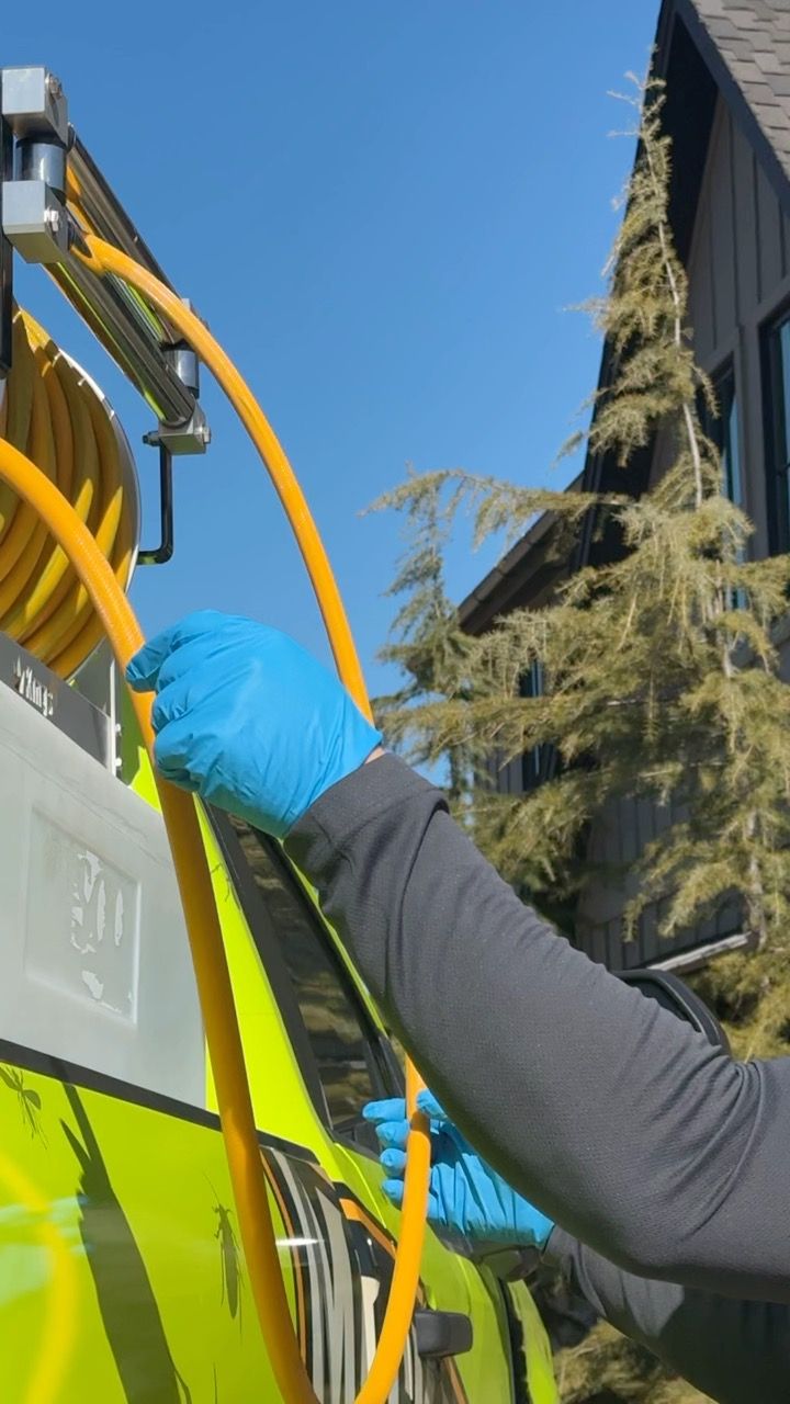 Person in blue gloves holding a yellow hose near a truck; house and trees in the background.