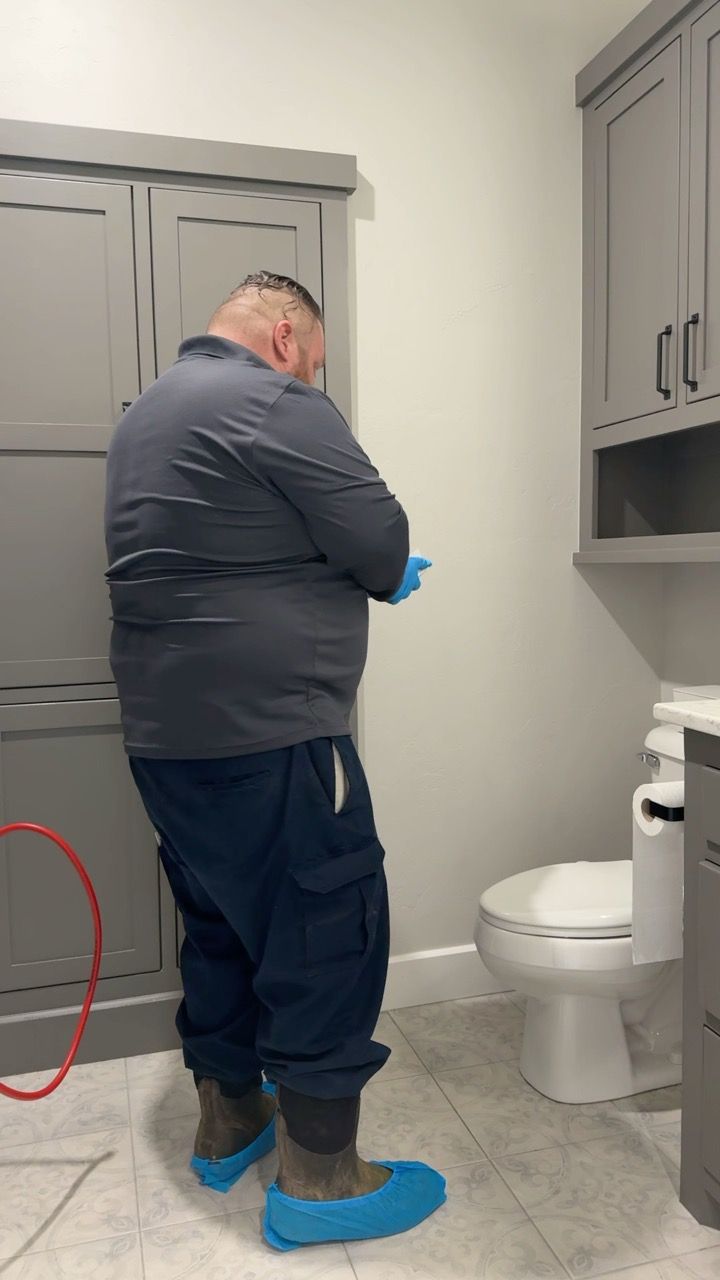 Person in blue scrubs standing near a toilet in a bathroom with gray cabinets and a red hose.