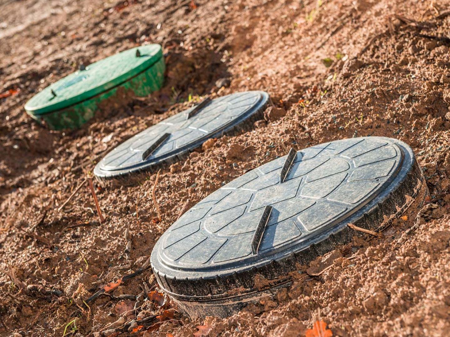 Three round utility covers on a dirt slope, with one green cover and two gray covers.