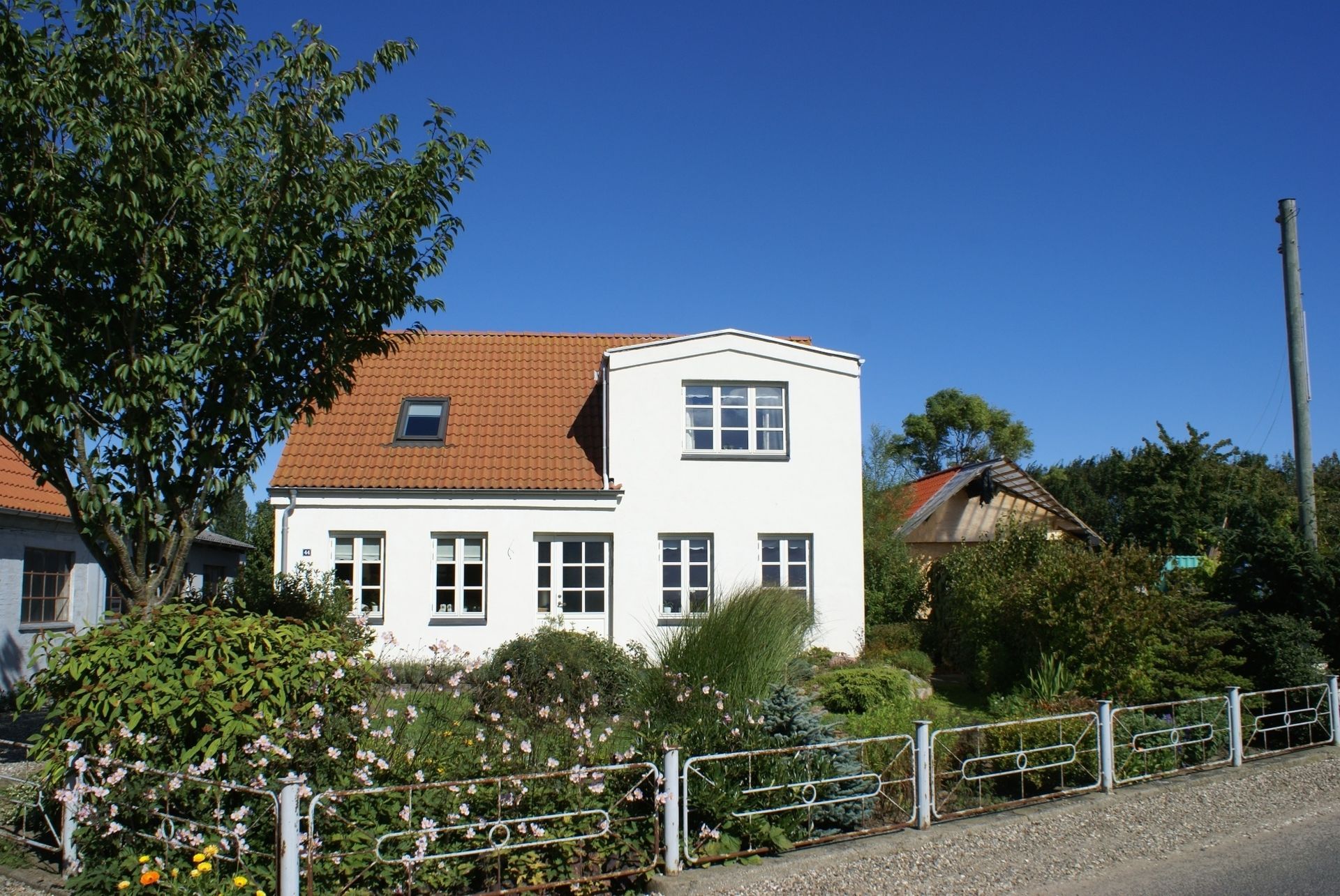 Hvidt hus med rødt tegltag, omgivet af grønt løv, blå himmel.