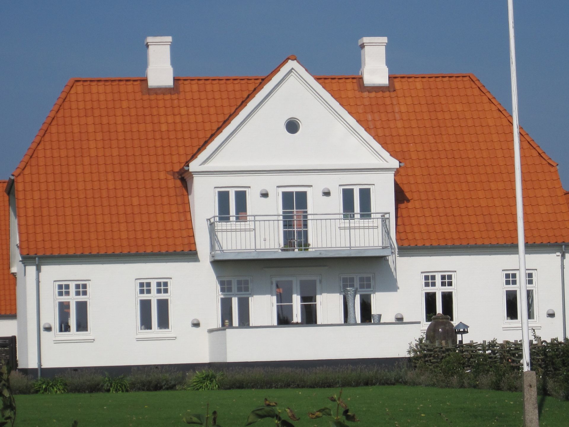 Hvidt hus med rødt tegltag, balkon og skorstene mod en blå himmel.