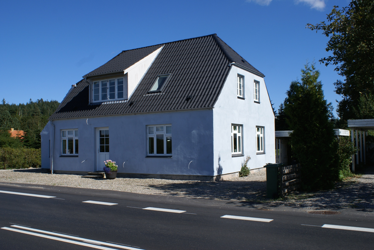 Lyseblåt hus med mørkt tag, ved siden af ​​en vej, under en klar blå himmel.