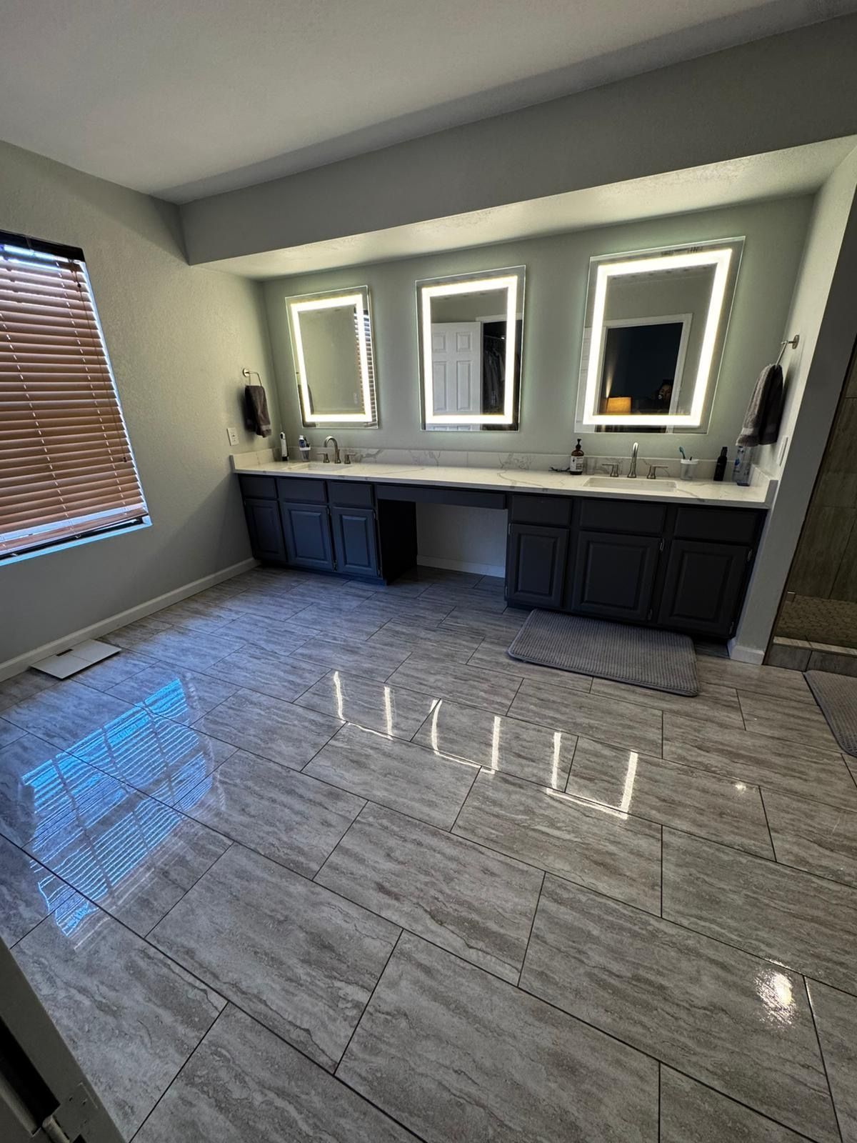 Bright bathroom with double vanity, three illuminated mirrors, dark cabinets, and glossy gray tile floor