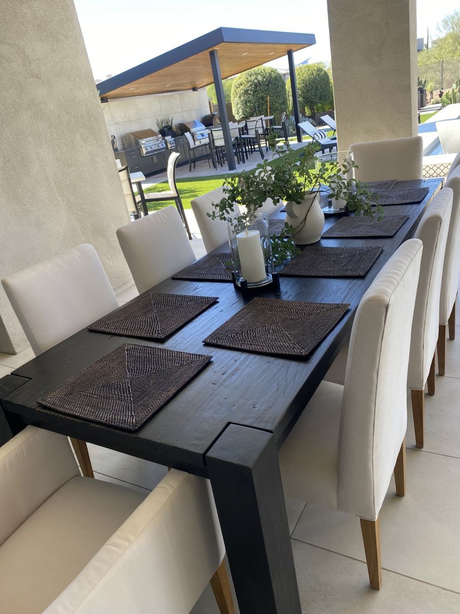 Outdoor dining table with white chairs, dark placemats, and potted plants under a covered patio