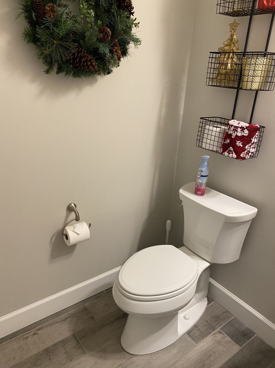 Small bathroom corner with a white toilet, toilet paper holder, wreath, and wire shelf with decor.