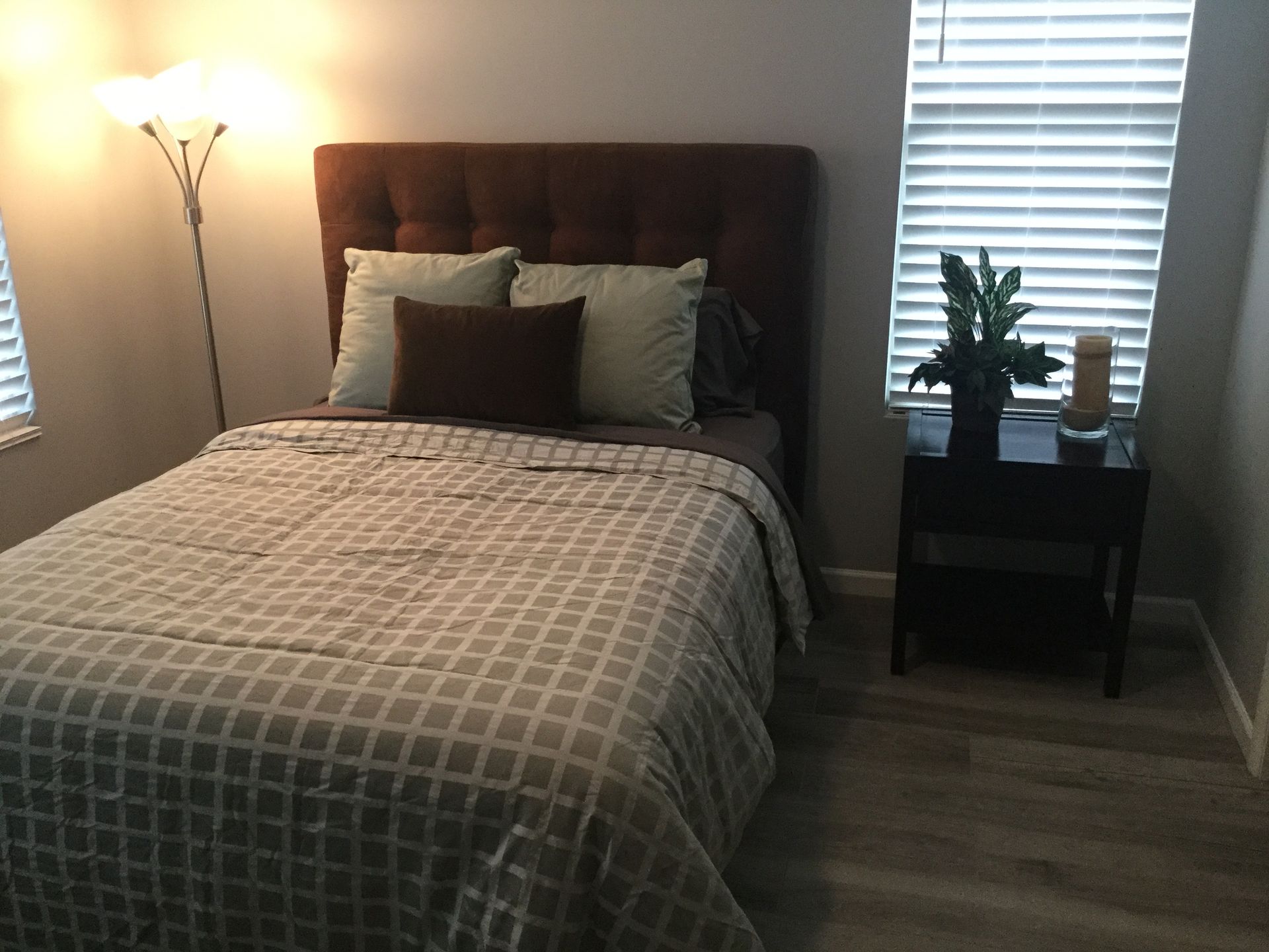 Small bedroom with a made bed, floor lamp, and window-side nightstand with a plant.