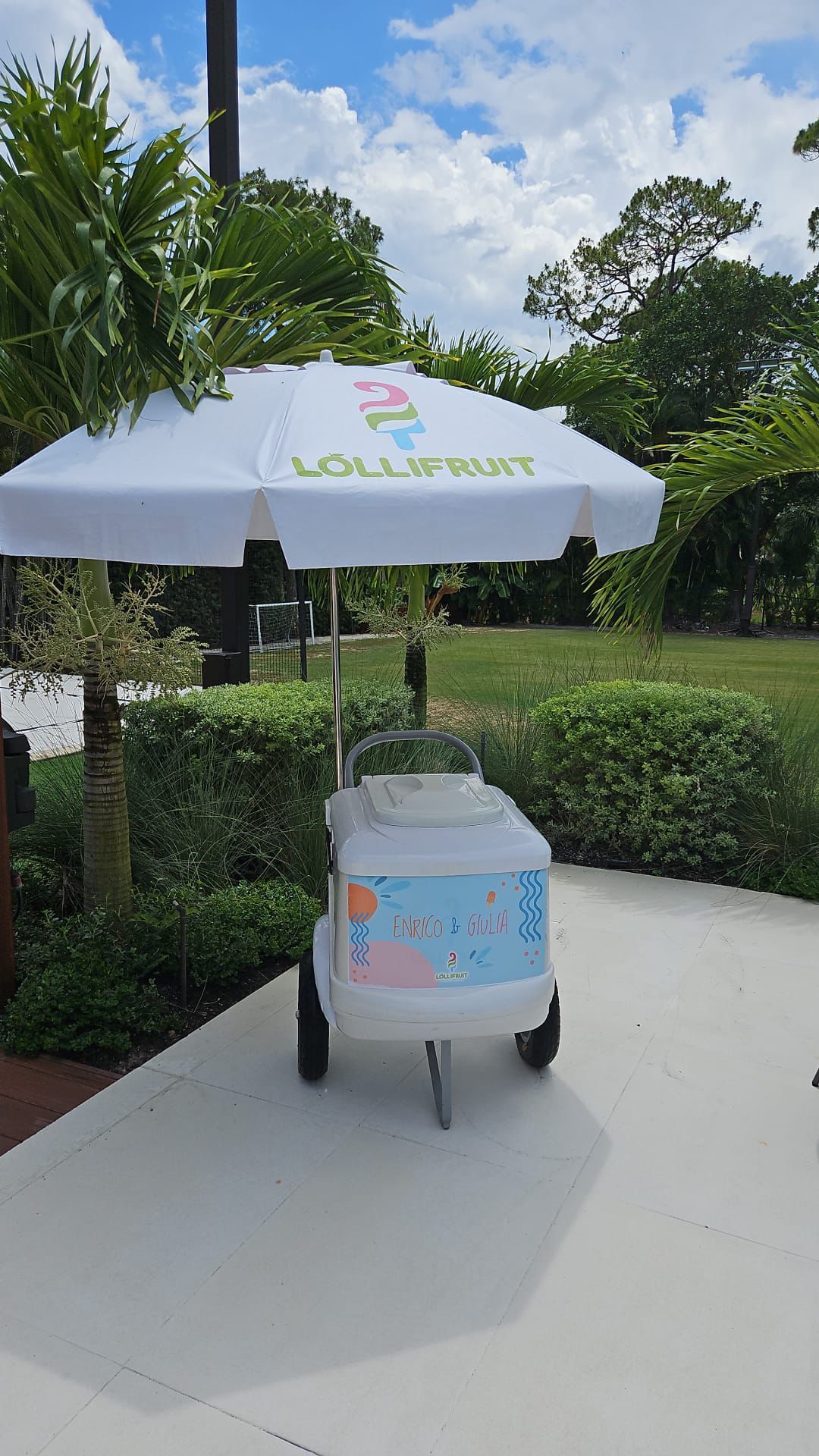 A white ice cream cart with a white umbrella is parked on a sidewalk.