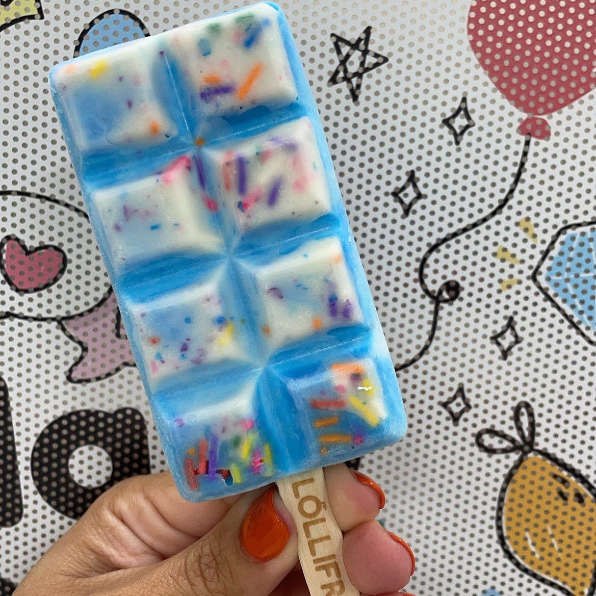 A person is holding a blue and white chocolate bar with sprinkles on a stick.