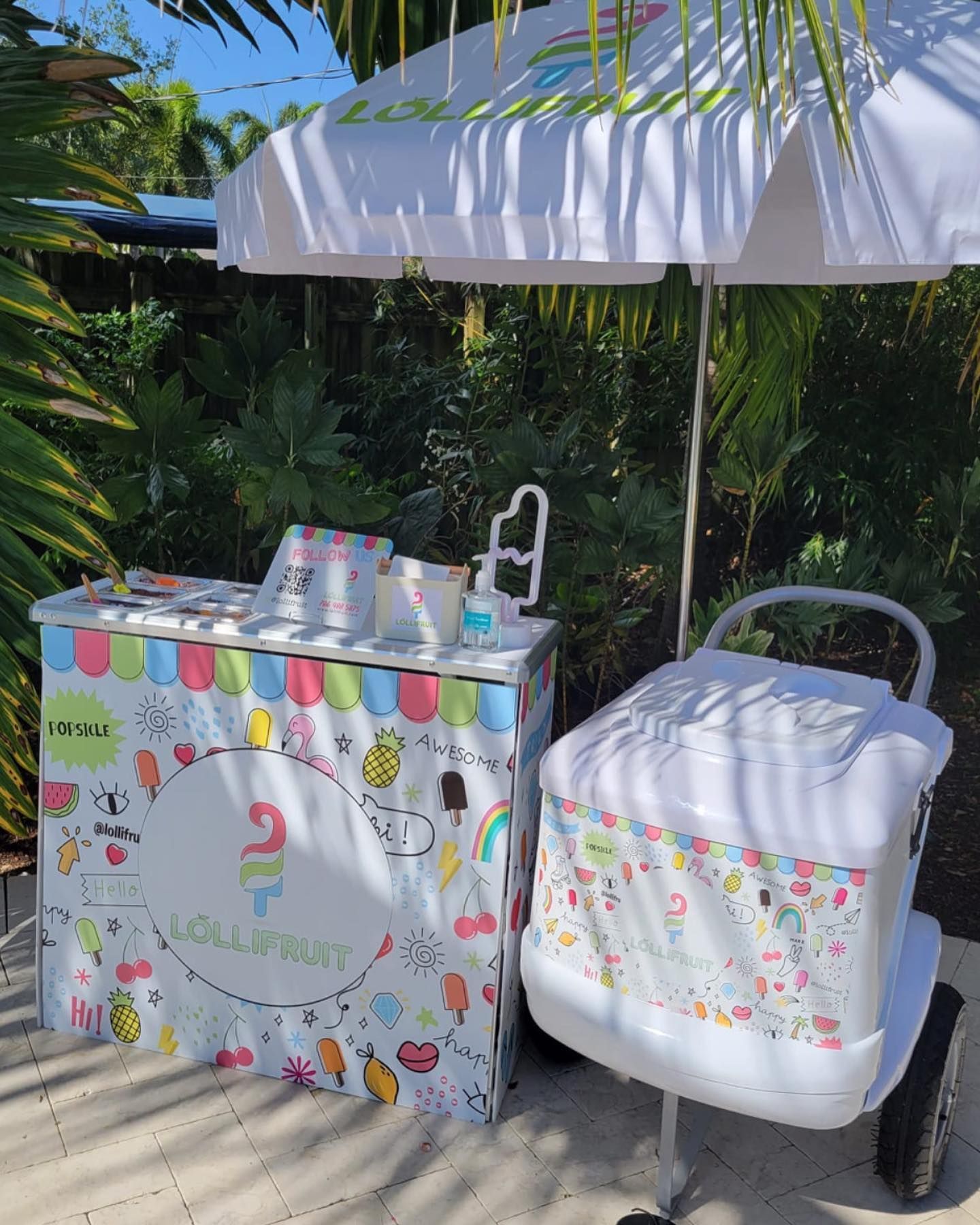 An ice cream cart is parked next to a cooler under an umbrella.