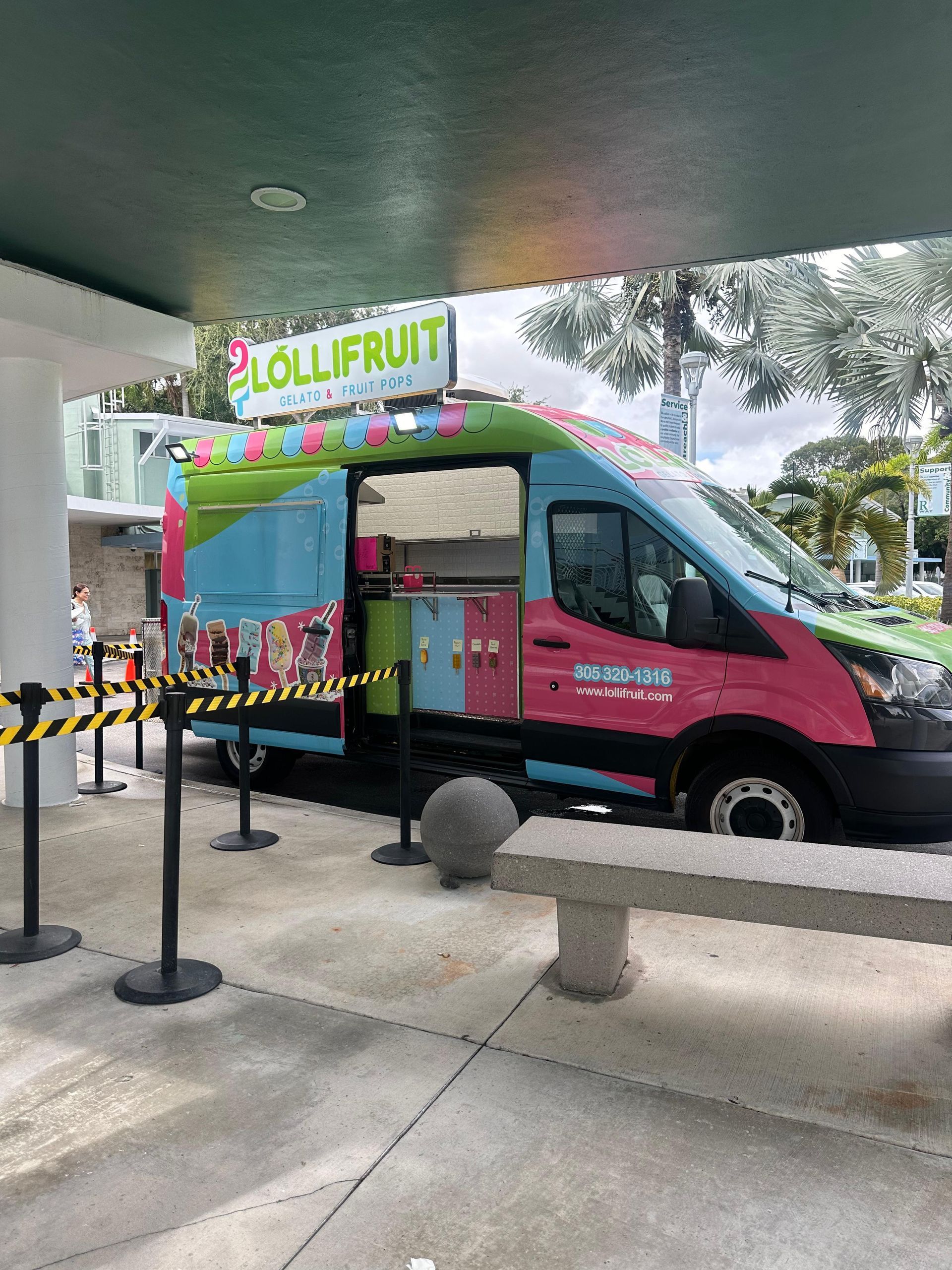 Colorful LolliFrut food truck parked under a canopy, with rope stanchions in front and trees nearby