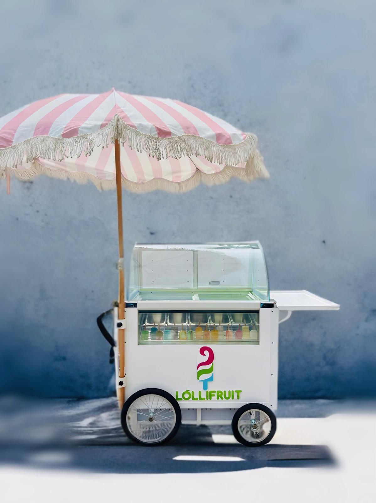 White and pastel pink ice cream cart with umbrella against a blue wall
