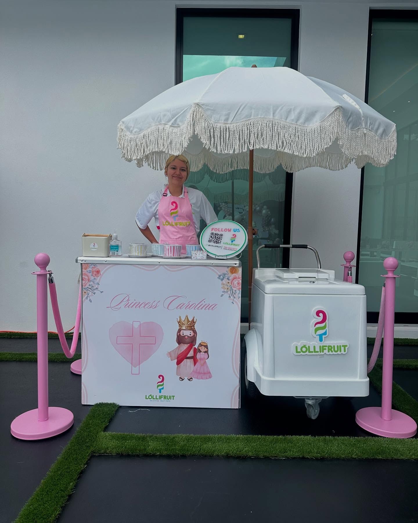Pink dessert cart with large umbrella and decorated sign outside a building
