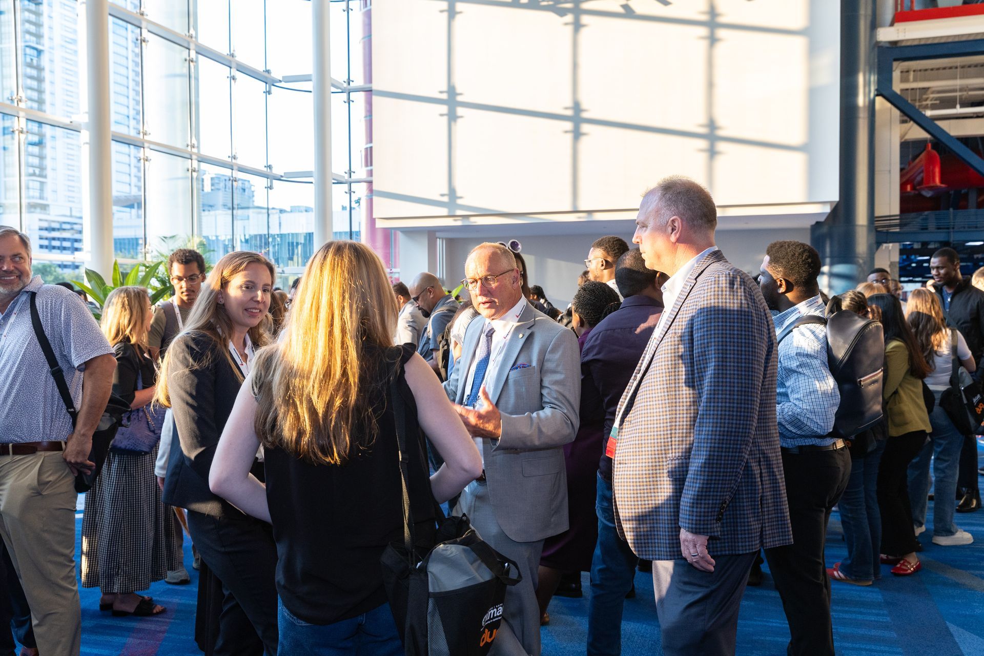 People in a large bright room with a glass wall, some are talking. Others walk and stand in small groups.