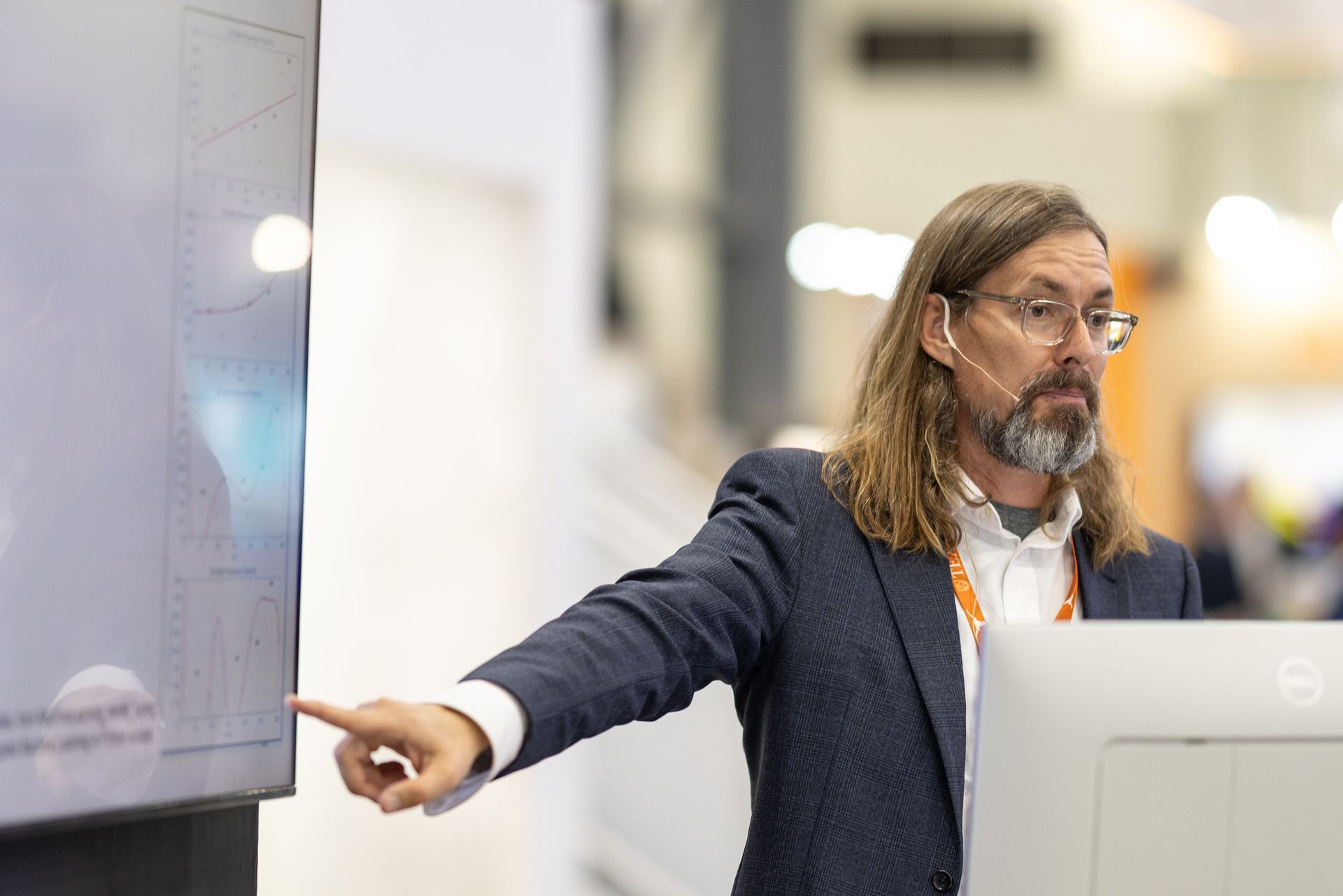 Man in blue blazer points at a graph on a screen, presenting data at a conference.