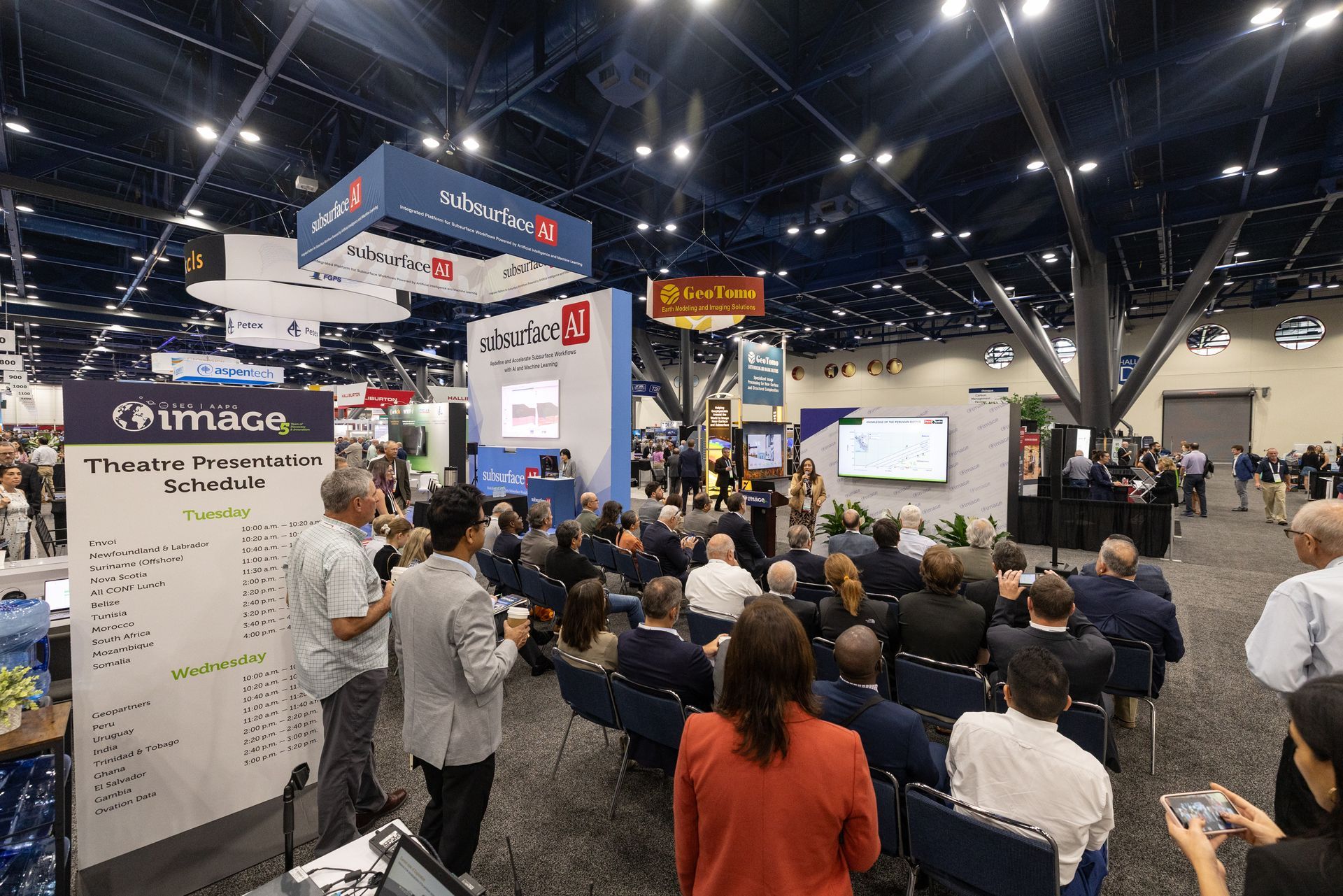 Conference attendees seated, watching a presentation at a trade show. Booths and displays surround them.