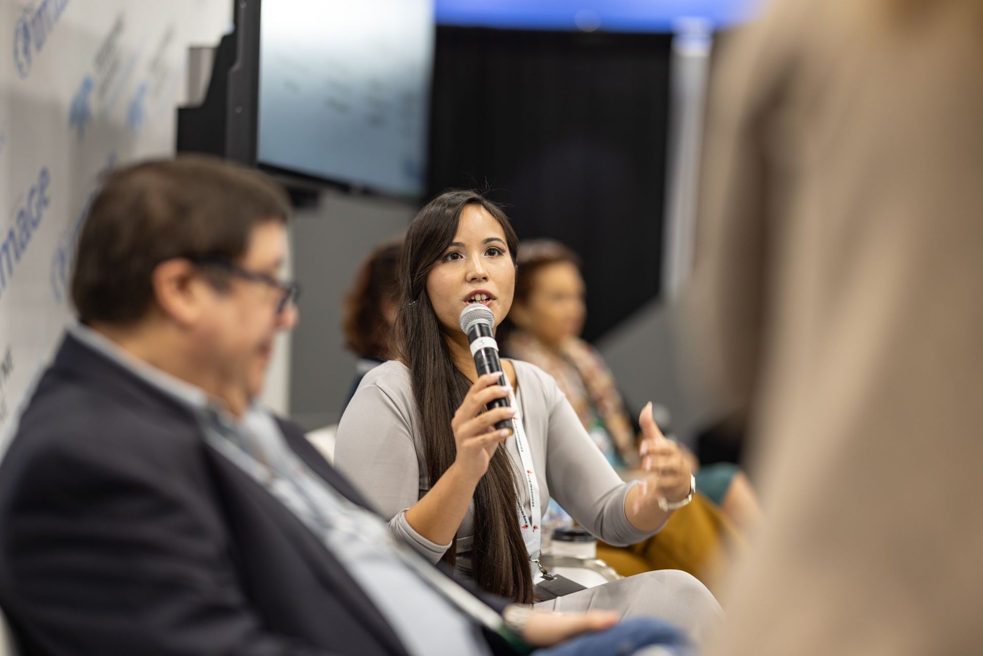 Woman speaking into a microphone during a panel discussion. Others are seated nearby.