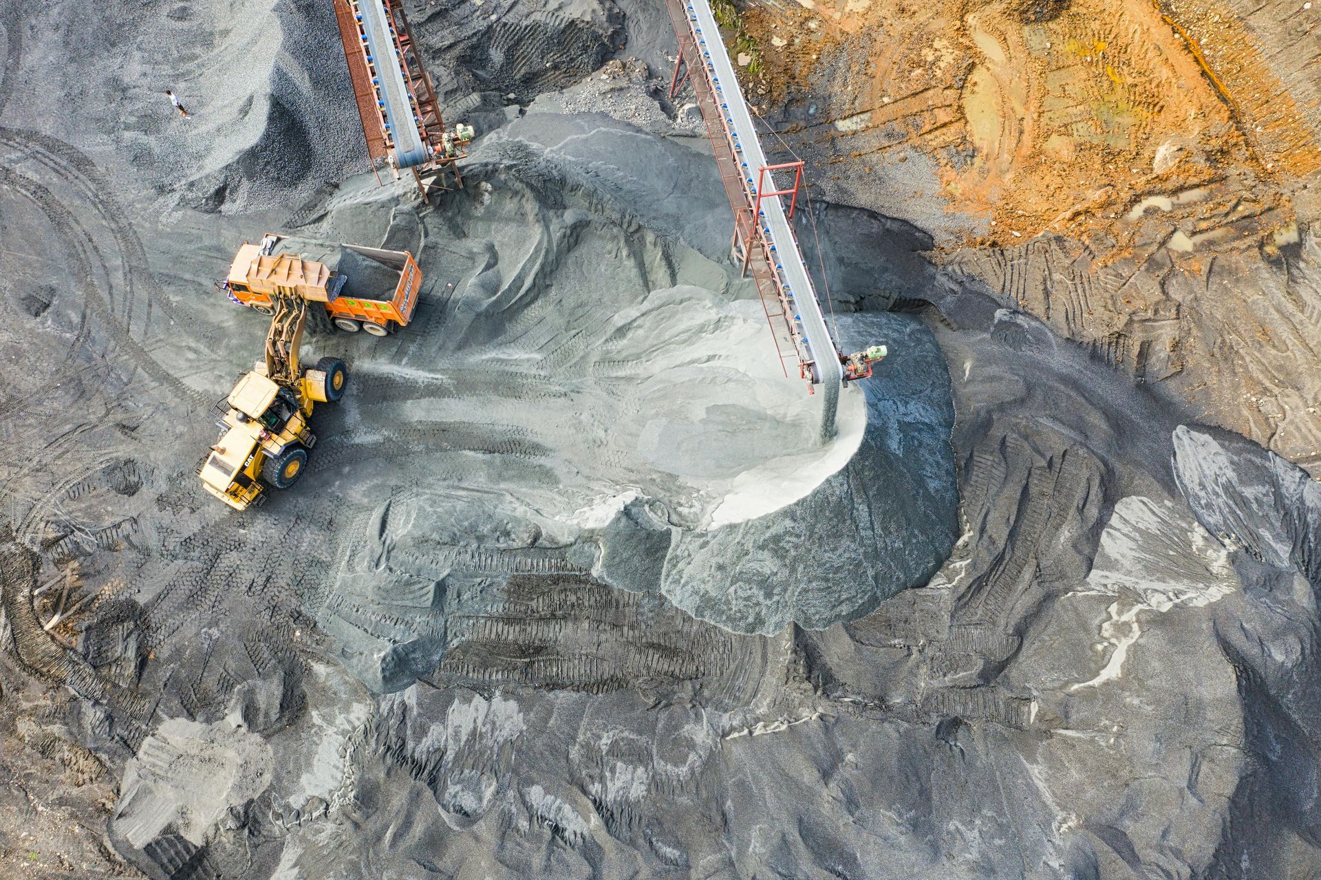 Overhead view of an excavator loading material onto a conveyor belt in a quarry, dusty environment.