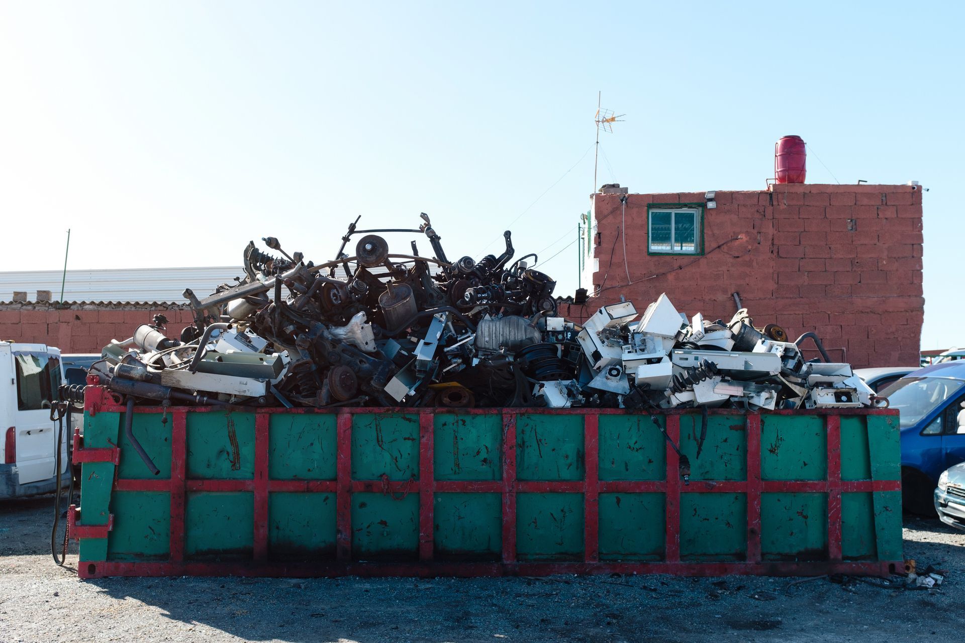 A large green dumpster filled with scrap metal in front of a brick building.