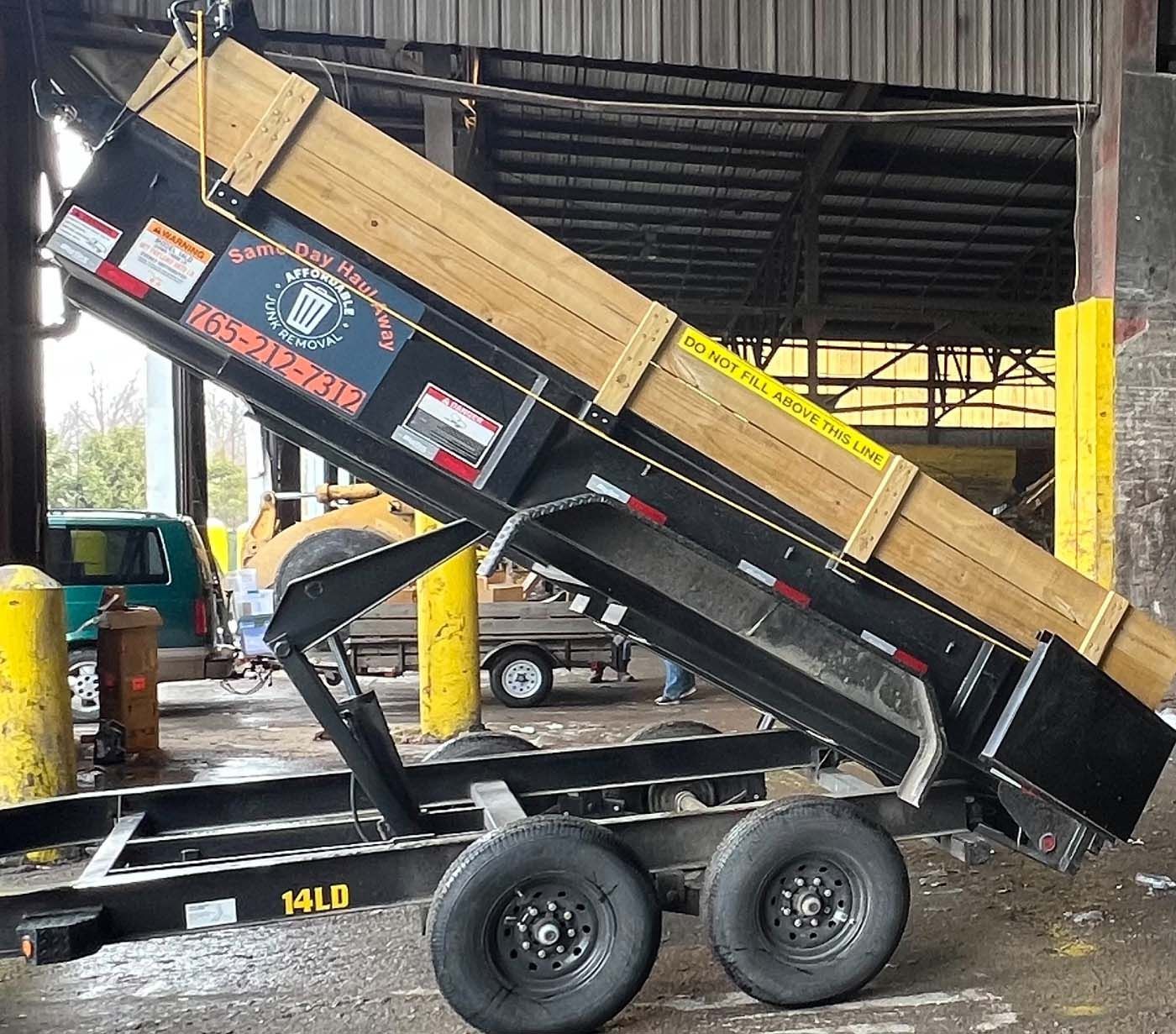 A black dump trailer with a wooden frame is raised. The trailer is parked inside a building.