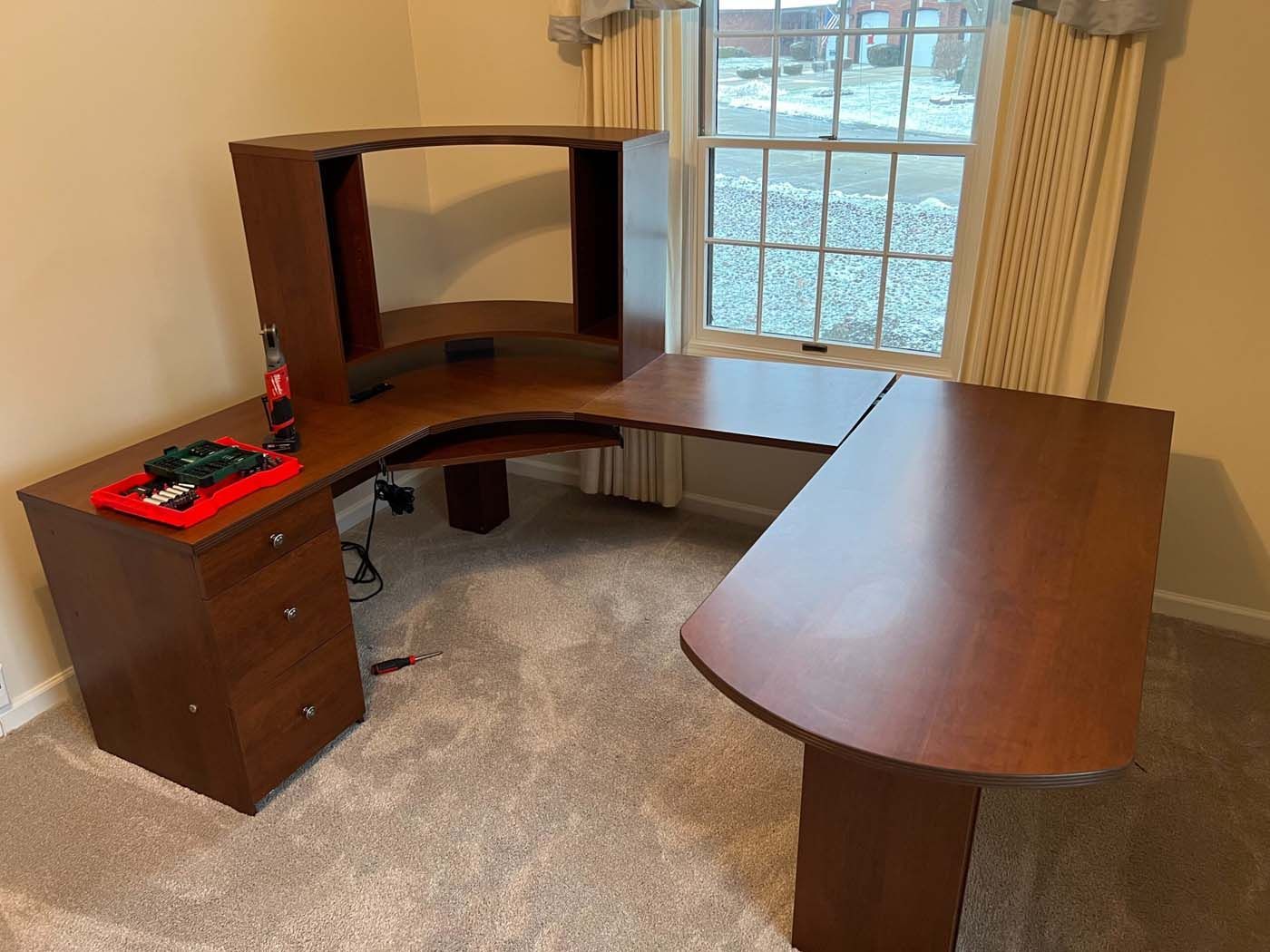 Brown corner desk with attached hutch, drawers, and side table in a room with a window and carpet.