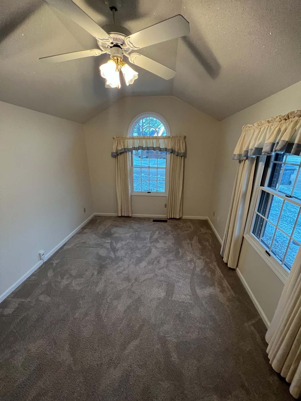 Empty room with brown carpet, two windows with curtains, and a ceiling fan.