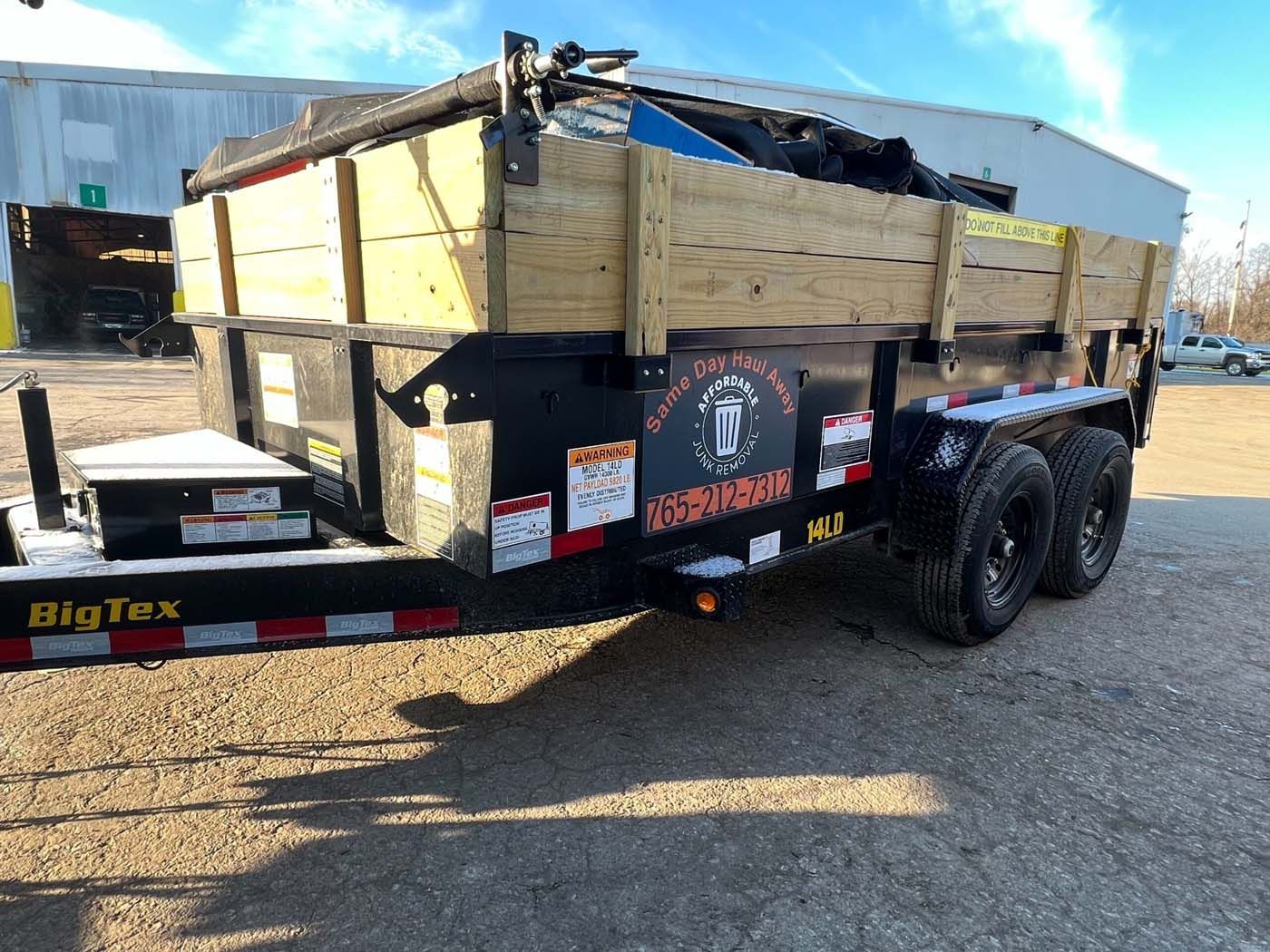 Black trailer with wooden sides, loaded with debris, parked outside.