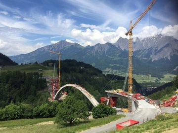 In den Bergen wird eine Brücke gebaut, im Vordergrund ist ein Kran zu sehen.