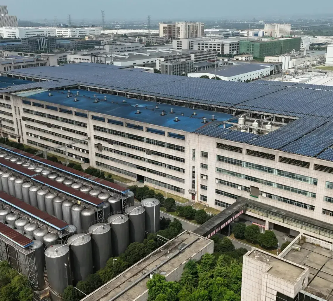 An aerial view of a large industrial factory complex with extensive rooftop solar panels and silver storage silos.