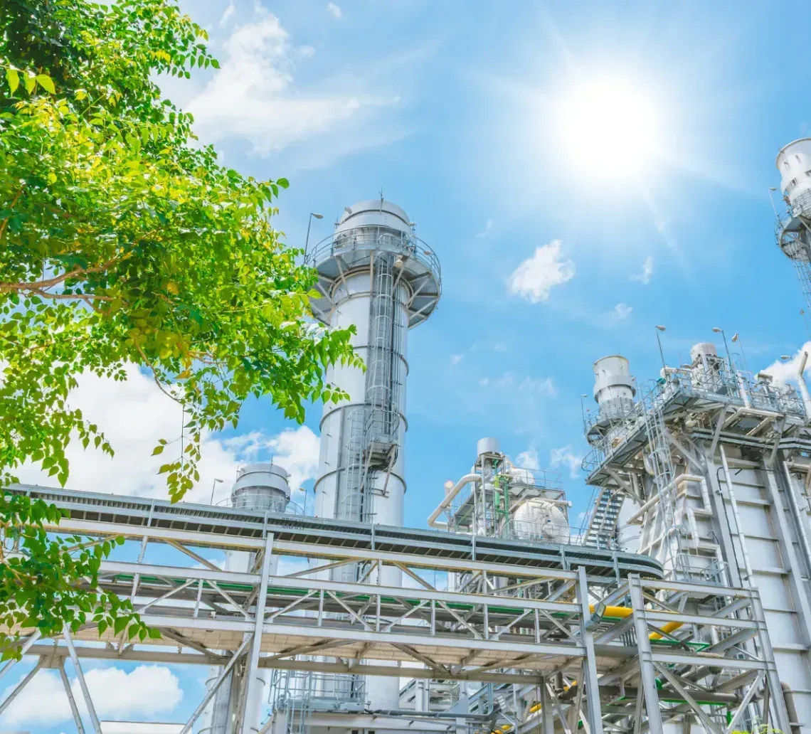 Industrial power plant with tall chimneys and pipe networks under a bright sun and clear blue sky, next to a green tree.