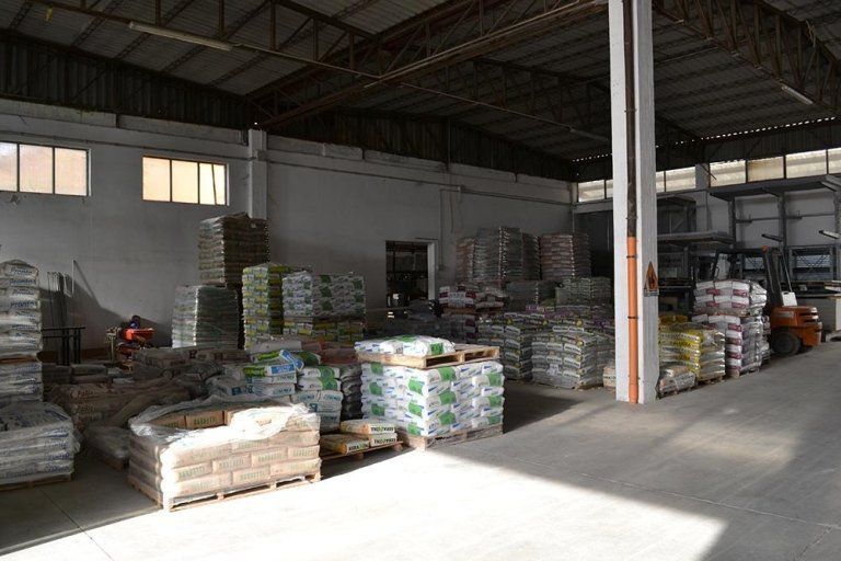 L'interno del magazzino è pieno di sacchi di materiali impilati su pallet. La luce del sole entra dalle finestre alte.