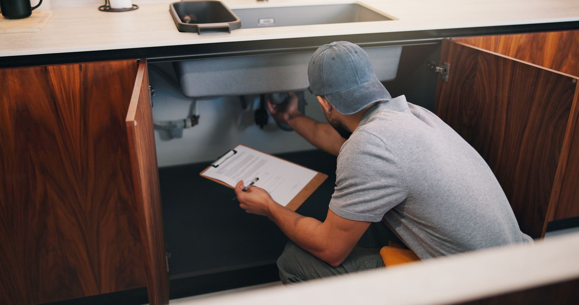 Man crouching under a desk, working on cables with a clipboard in a wood-paneled office cubicle.