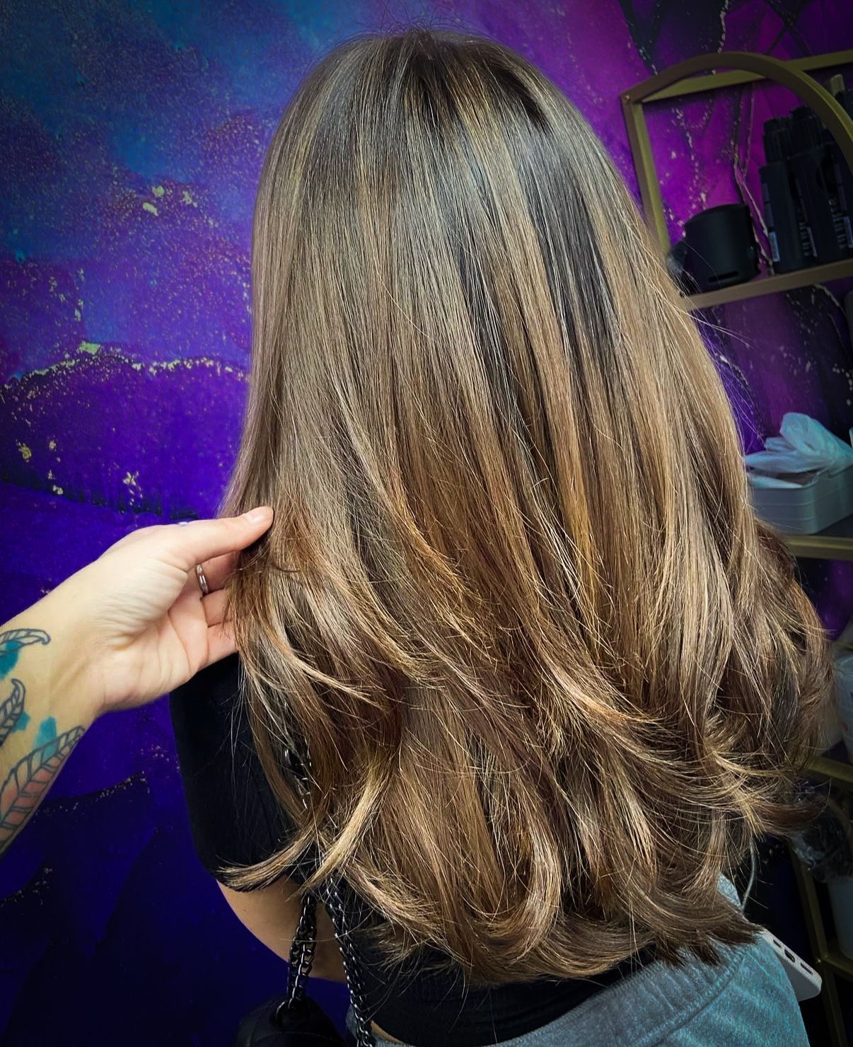Hand touching layered brown hair with highlights in front of a purple backdrop.