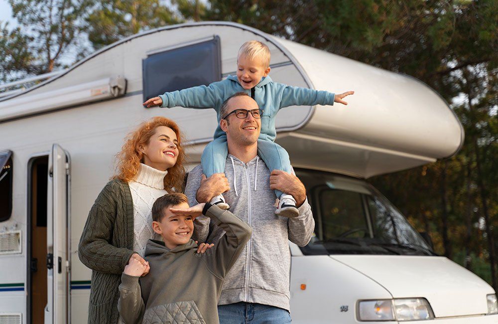 Family of four smiles outdoors, with children playing on parents' shoulders near a camper van.