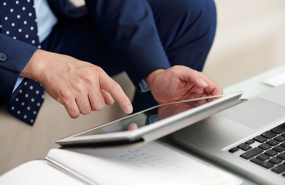 A person in a suit uses a tablet while seated next to a laptop and notepad.