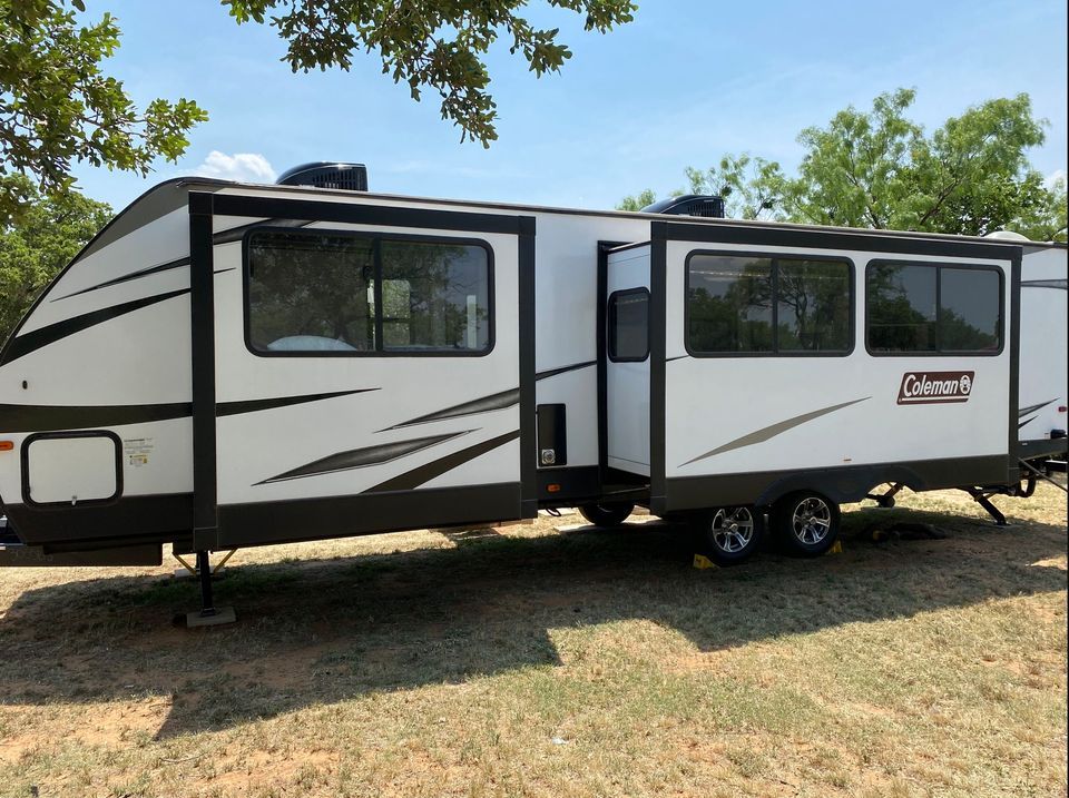 A white and black Coleman travel trailer parked in a grassy area under a clear blue sky.