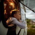 Boy with short hair leans out of an RV window, gazing pensively at a scenic landscape.