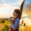 Woman in a vest gazes thoughtfully outdoors, holding a cup, with sunset and fields in background.