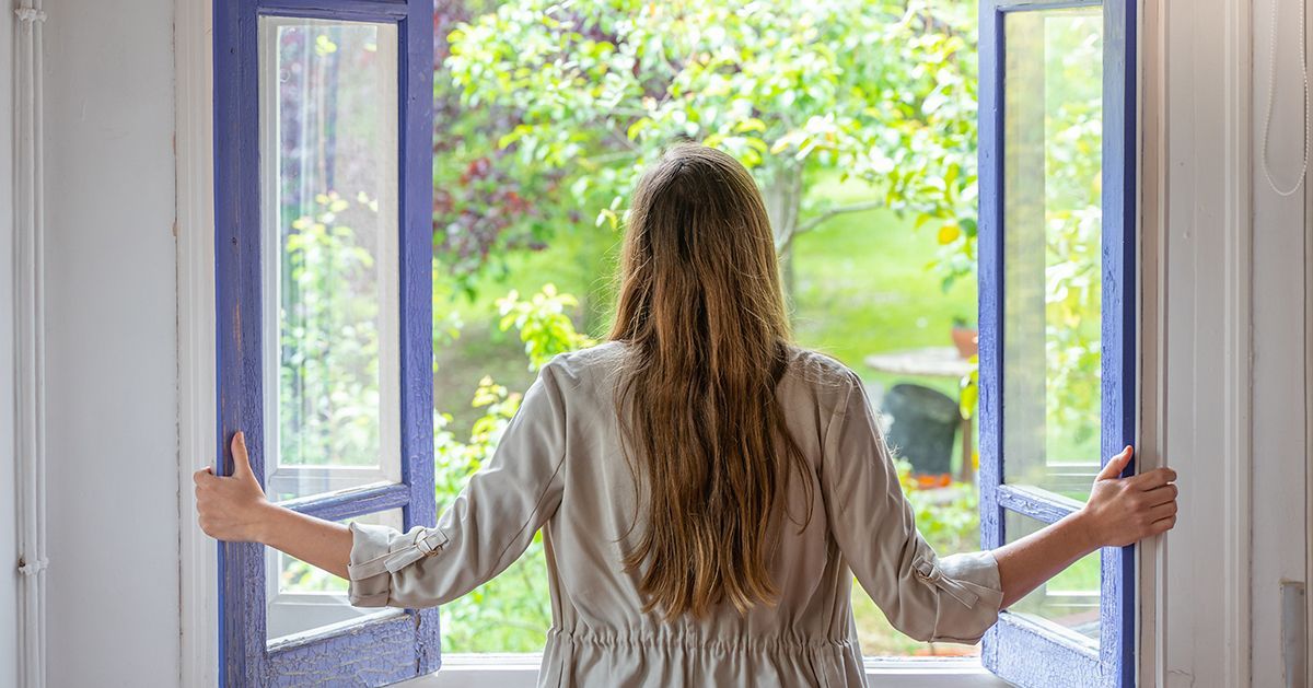 Eine Frau steht mit ausgestreckten Armen vor einem offenen Fenster.