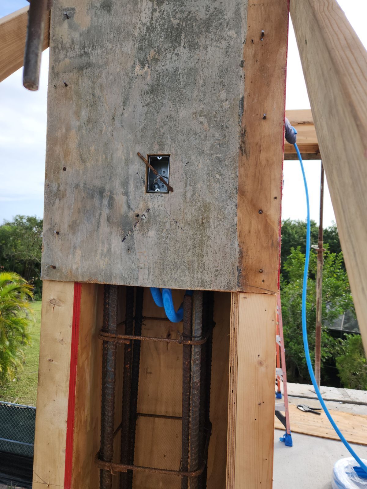Construction of a concrete column with rebar visible inside wooden formwork. Blue conduit extends downwards.